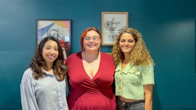 Three women standing side by side, smiling with art hung on a wall behind them.