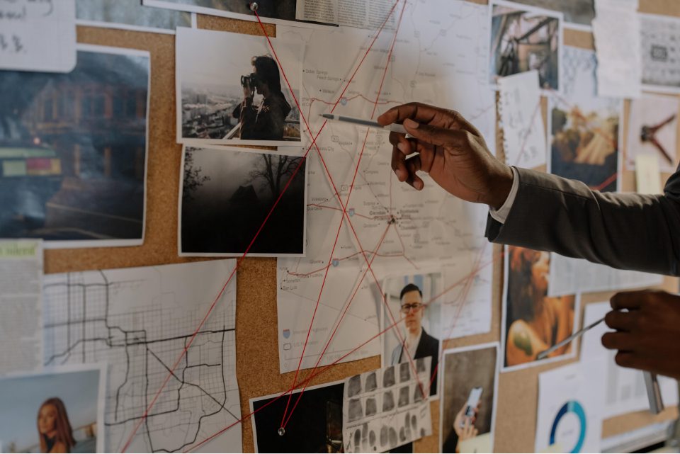 Investigator pointing at evidence board filled with photos, maps, and headshots connected with red string.