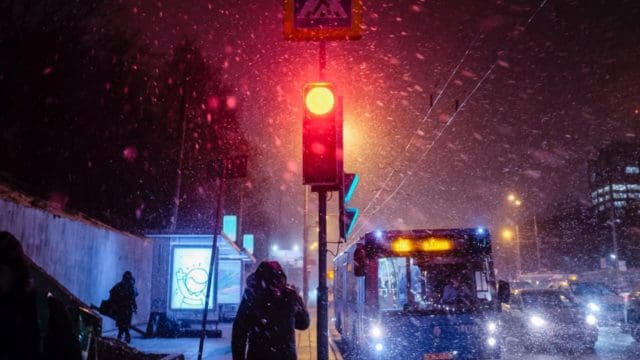 Crowded street with buses and cars. Snow is falling. There is a red traffic light glowing in the middle of the image.
