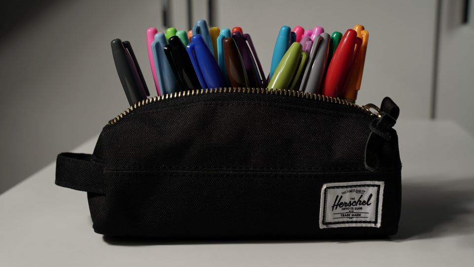 Black pencil bag, unzipped, filled with different colored sharpie markers sitting on a white table.
