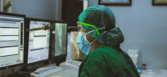 Doctor in green scrubs looking at multiple computer screens with sensitive medical information on them