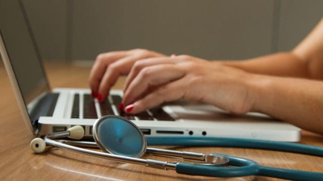 Female doctor typing on a laptop with a stethoscope on the desk.
