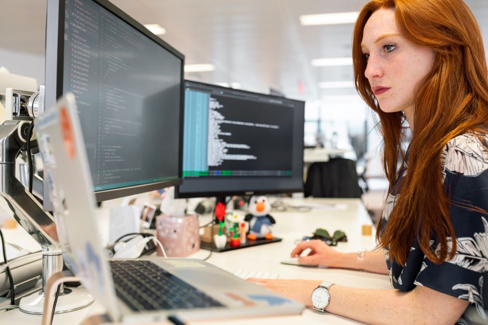 Female software developer with red hair looking at code on three monitors.