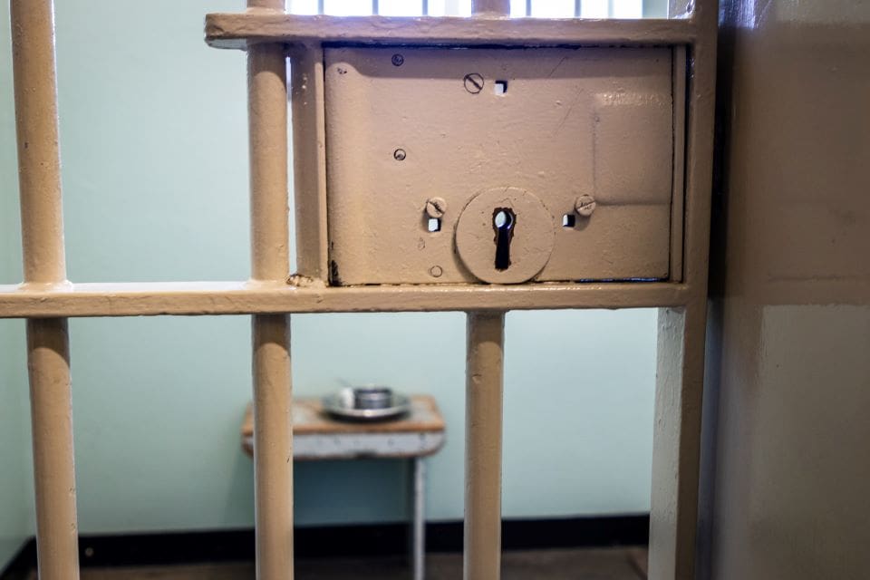 View into prison cell through metal bar door with key lock.